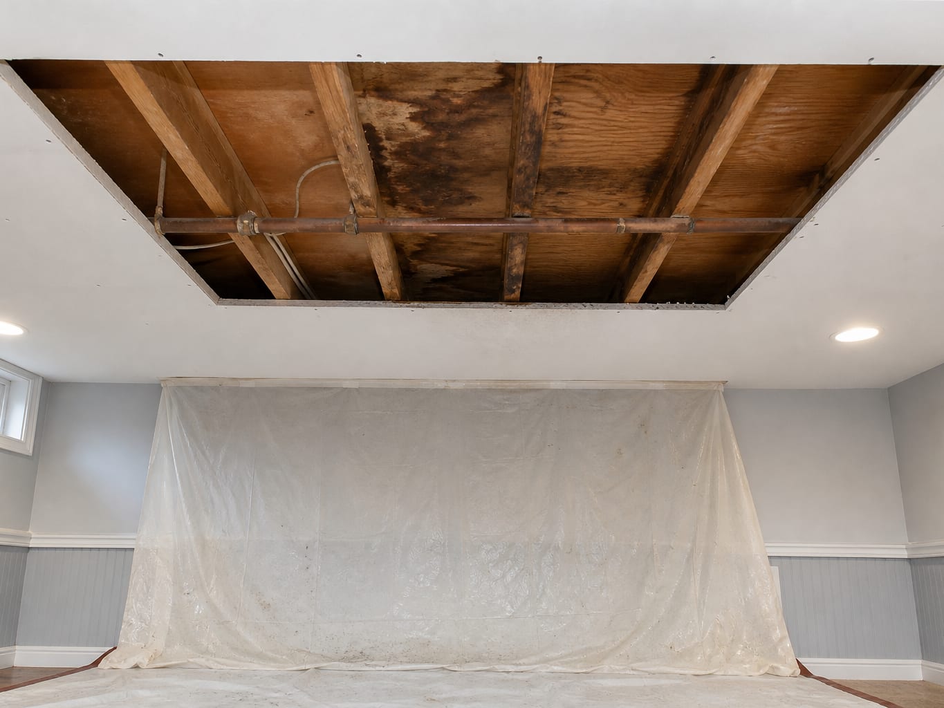 A wider view of a room where a section of ceiling drywall has been removed to expose the joists and the underside of the floor above, with staining that marks the path water took between levels