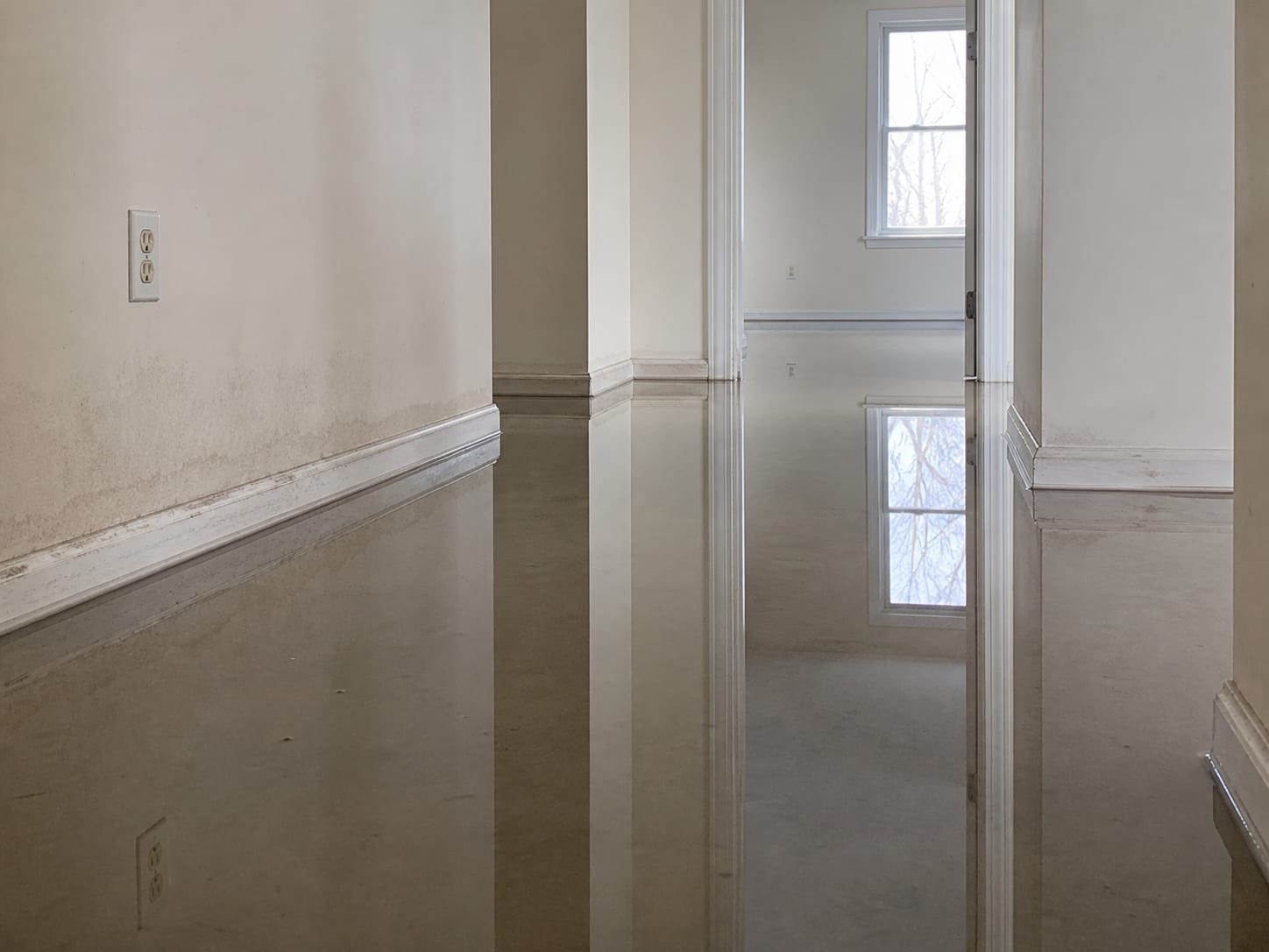 Standing water spreading across a ground floor hallway with an electrical outlet visible low on the wall just above the water line, no equipment running, in Plainsboro, New Jersey