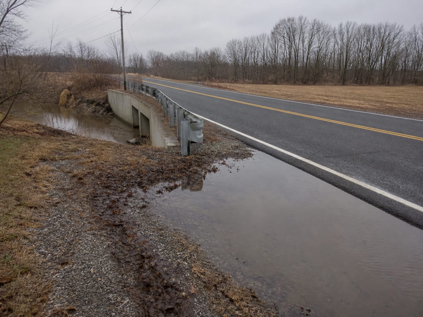 A low lying two lane road crossing in West Windsor, New Jersey with a concrete culvert headwall and standing water across the shoulder after rain