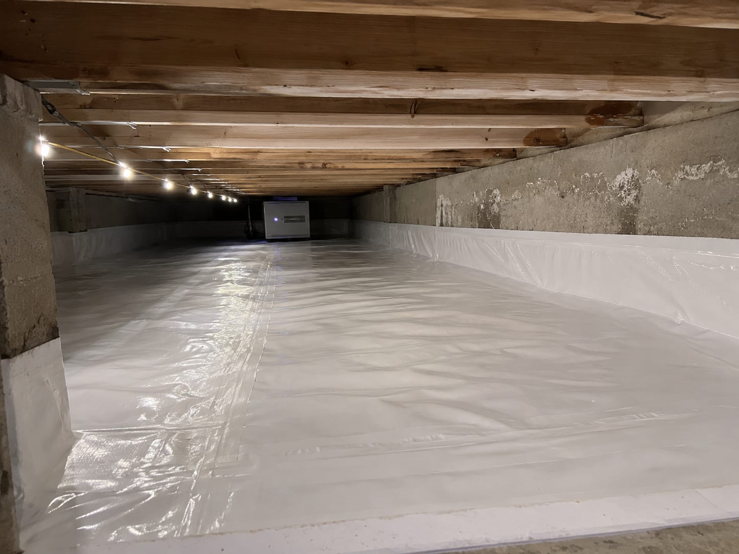 Crawl space under a New Jersey colonial home after mold remediation showing white vapor barrier encapsulation covering the ground and lower foundation walls with a small dehumidifier operating in the background