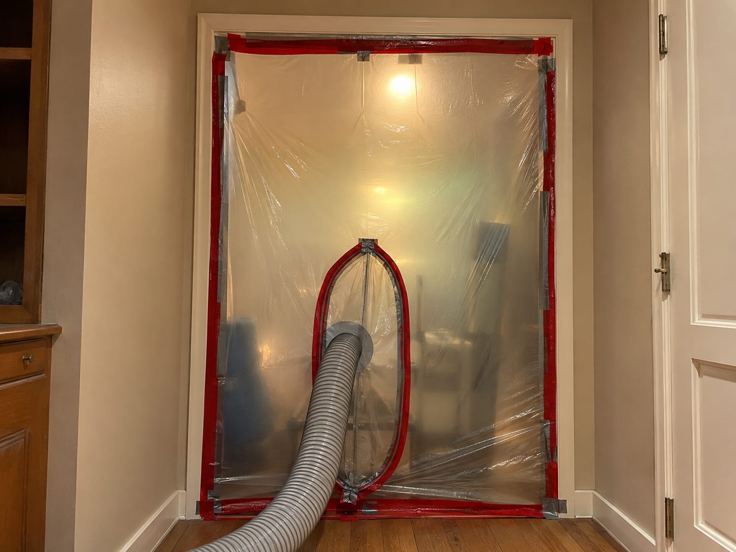 Clear plastic poly sheeting containment barrier sealed with tape across a doorway in a residential hallway, with a HEPA air scrubber intake hose passing through a zipper entry port