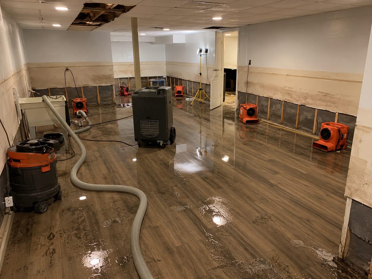 Flooded finished basement with standing water on laminate flooring, industrial extraction equipment positioned near baseboards, dehumidifiers and air movers running along walls