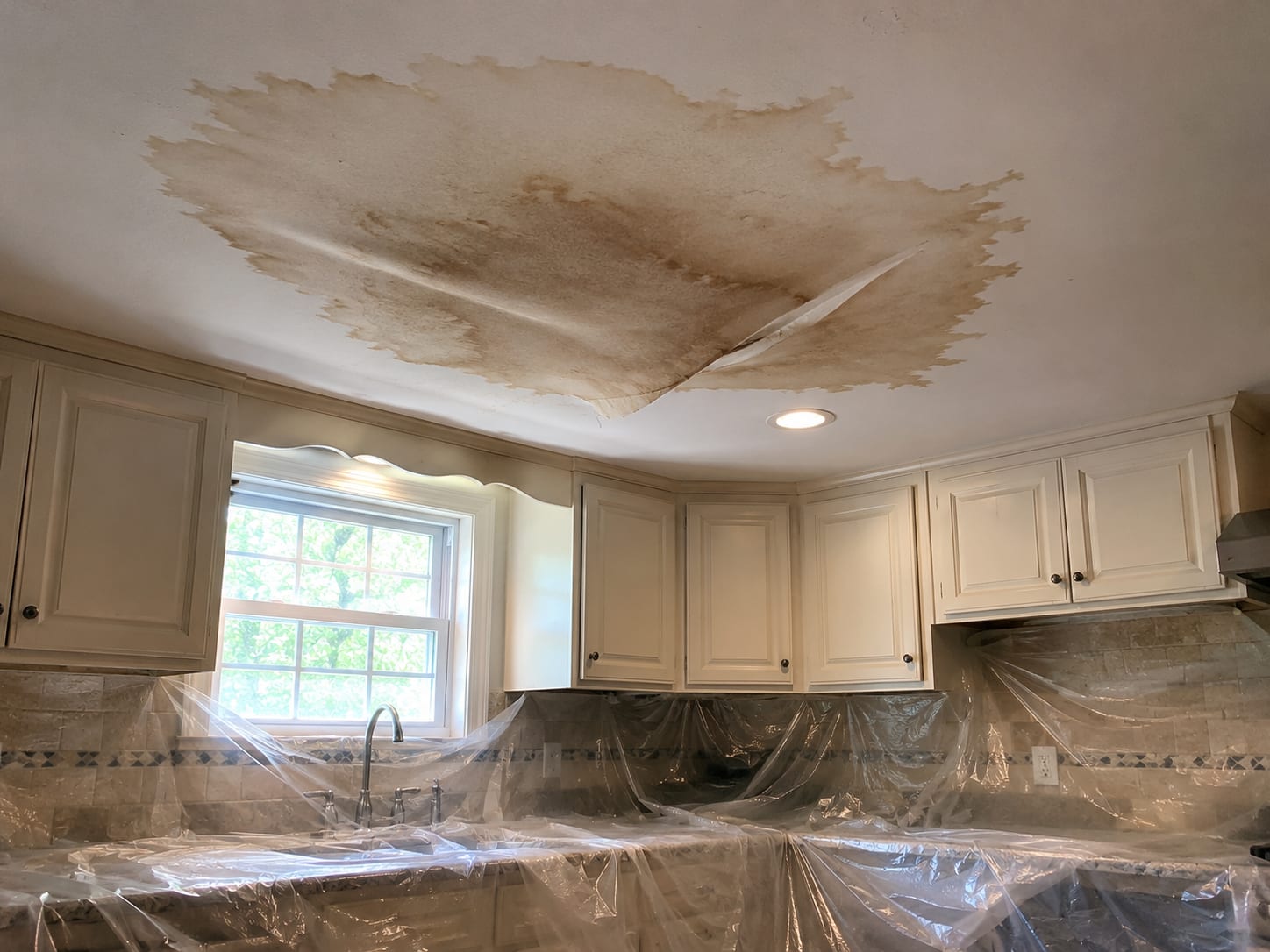 Water-damaged kitchen ceiling with sagging drywall, brown water stain spreading from a central point, and a plastic containment sheet spread across kitchen counters and floor below