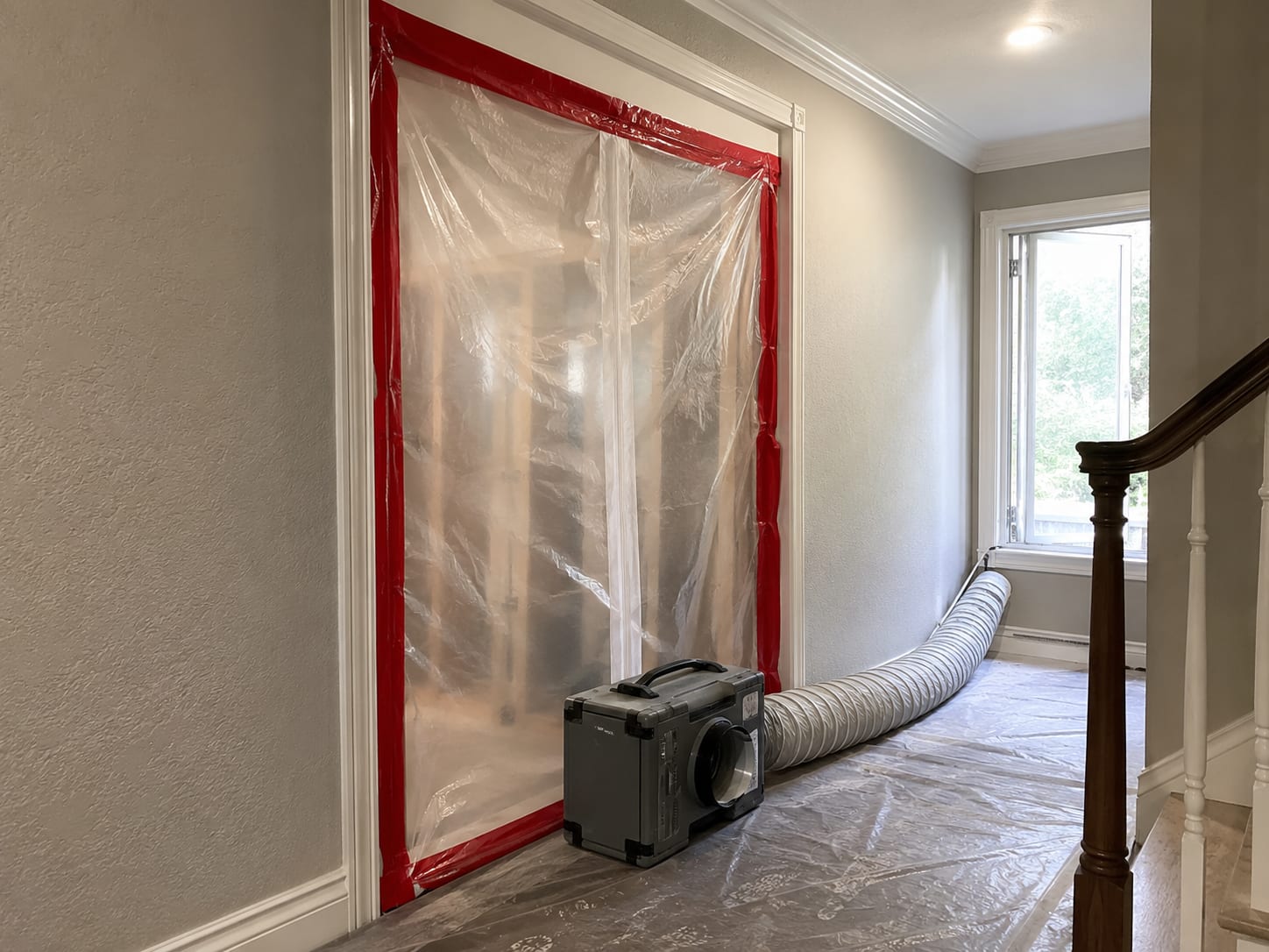 Active mold remediation containment zone inside a New Jersey home hallway showing heavy-duty plastic sheeting sealed across a doorway with a negative air machine exhausting outside through a window