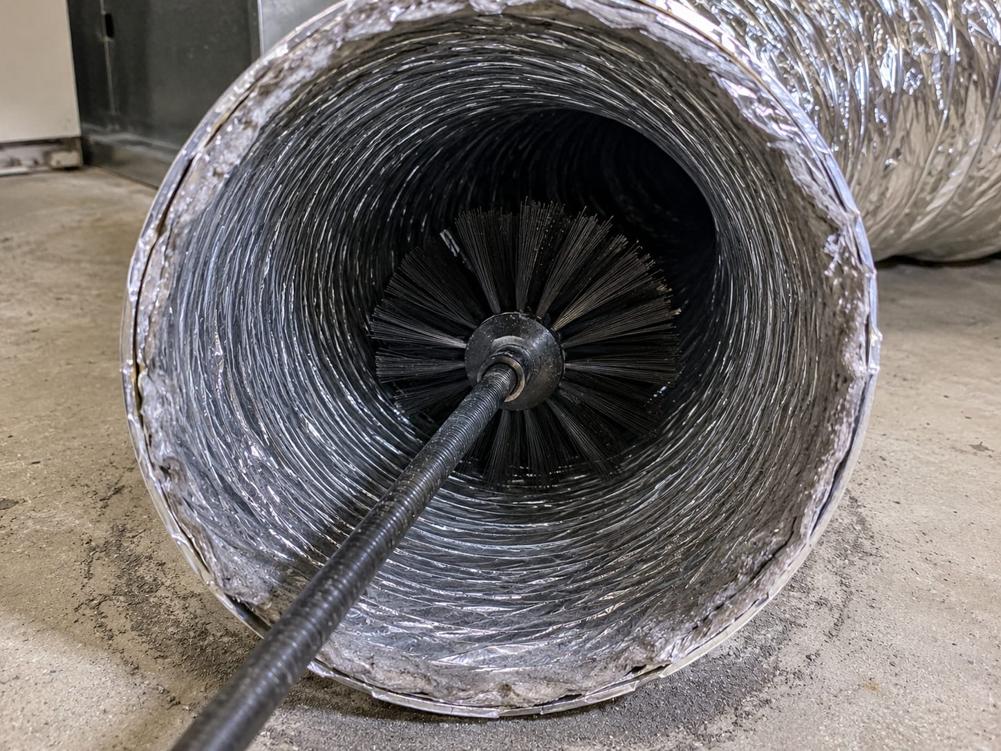 Interior perspective looking down a clean round flex duct with a rotary cleaning brush head visible partway inside, duct walls showing bare corrugated insulation and no debris