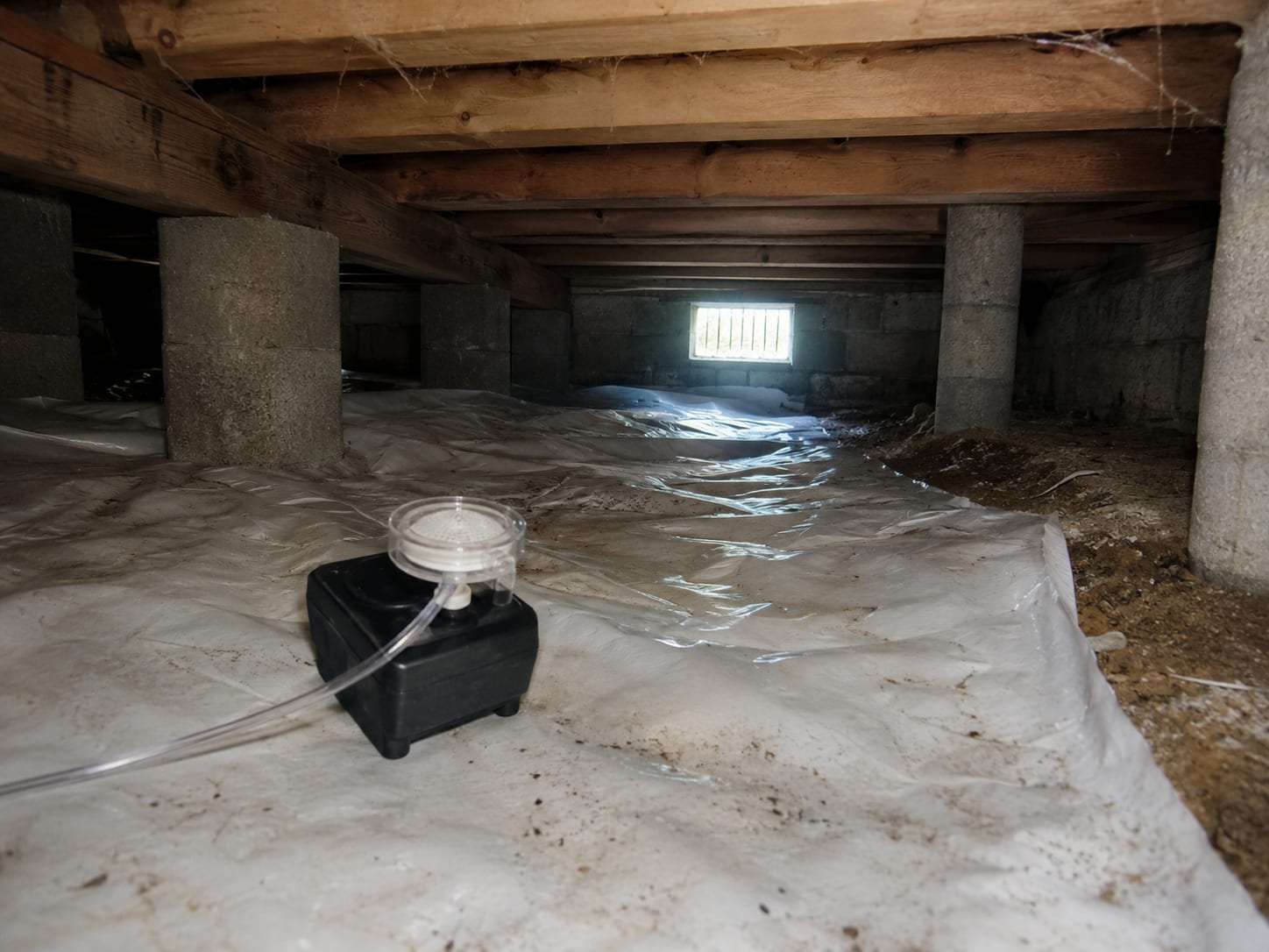 Air sampling pump resting on a vapor barrier in a low crawl space with concrete piers and dim natural light
