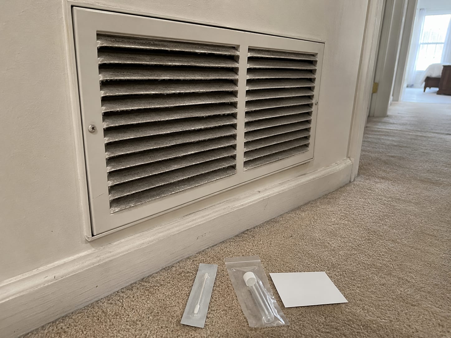 Surface swab collection kit beside a wall-mounted HVAC return air vent with accumulated dust on the grille louvers in a New Jersey home hallway