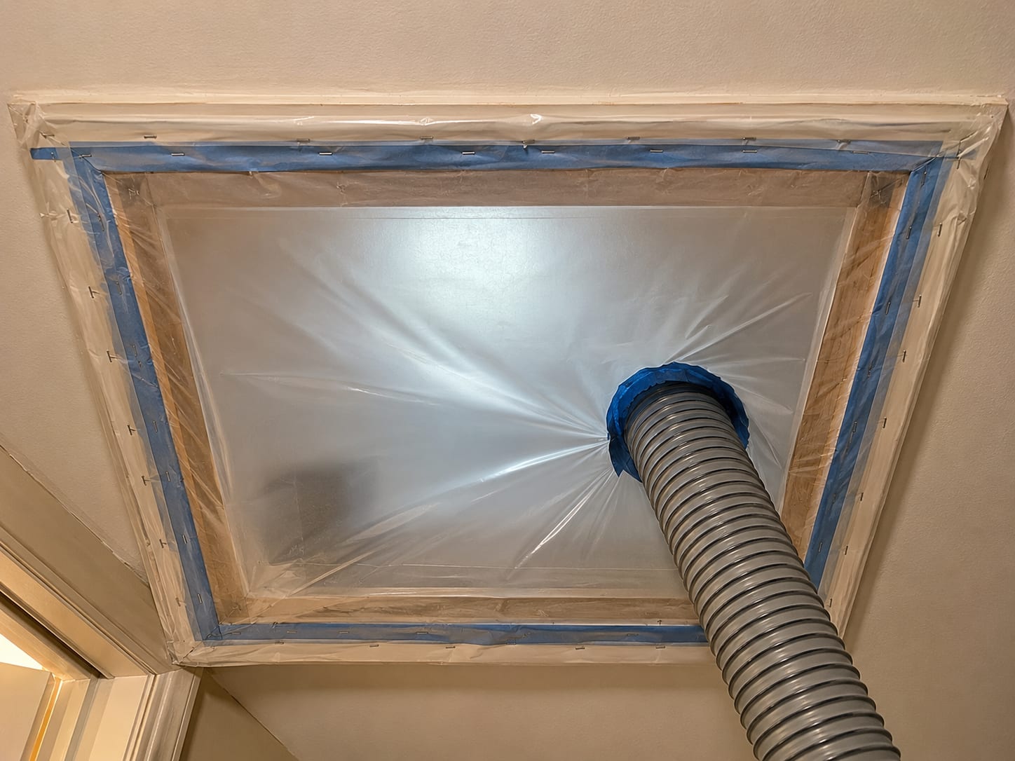 Close view of heavy poly containment sheeting stapled across attic hatch opening with a HEPA negative air machine exhaust hose exiting through a vent