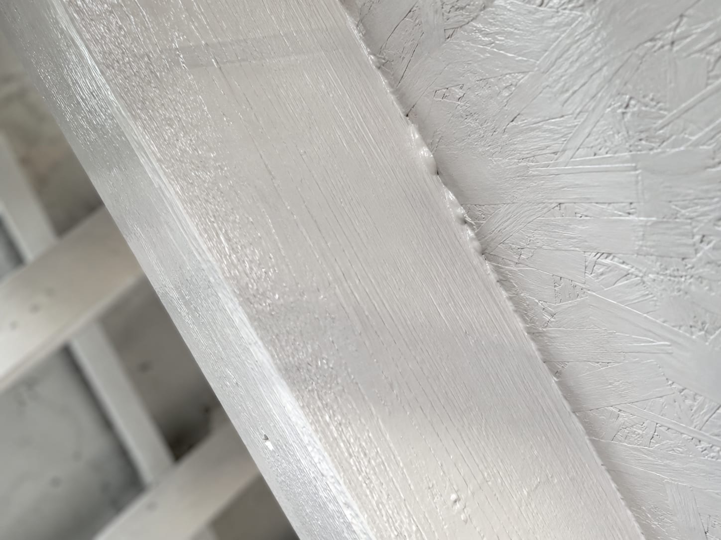 Macro close-up of a single dimensional lumber rafter and adjacent OSB sheathing panel fully coated in smooth white antimicrobial encapsulant with visible brush stroke texture