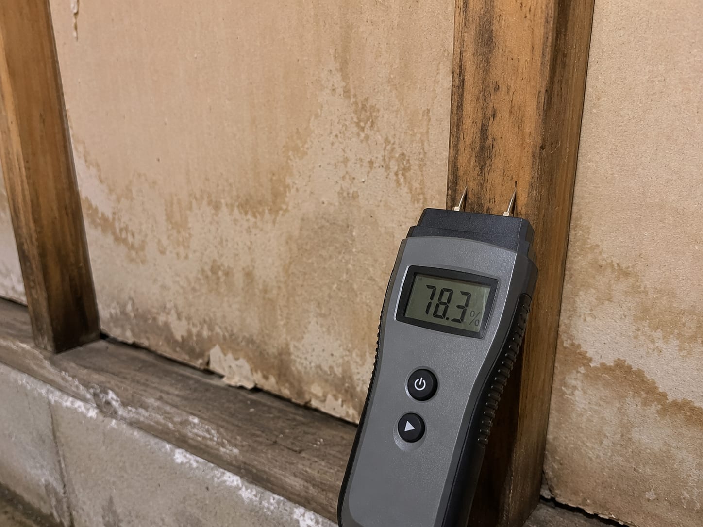 Digital moisture meter probe pressed against a damp basement wall stud showing elevated moisture reading on its small display