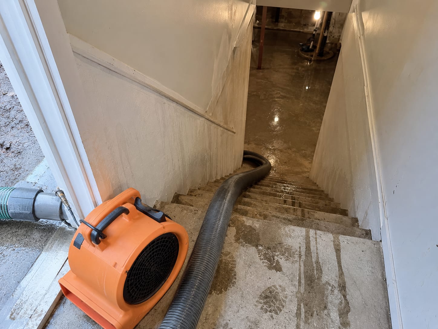 Industrial truck-mount extraction vacuum hose running into a basement entrance alongside an orange air mover blower
