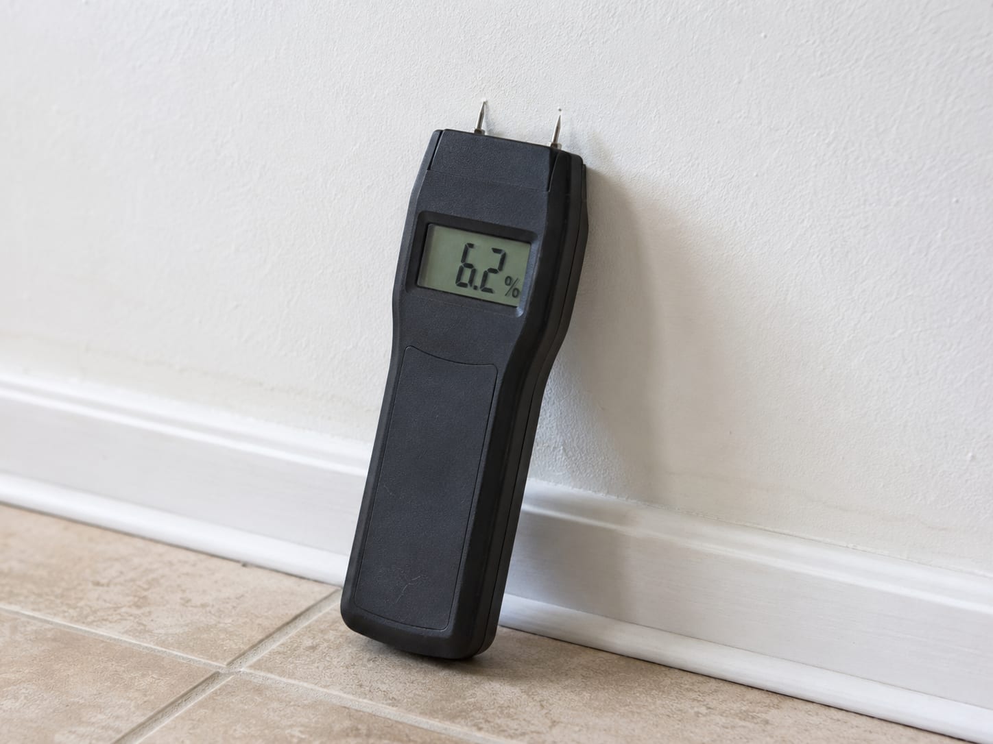 Digital pin-type moisture meter probe pressed against a remediated bathroom drywall surface displaying a low moisture reading after mold remediation