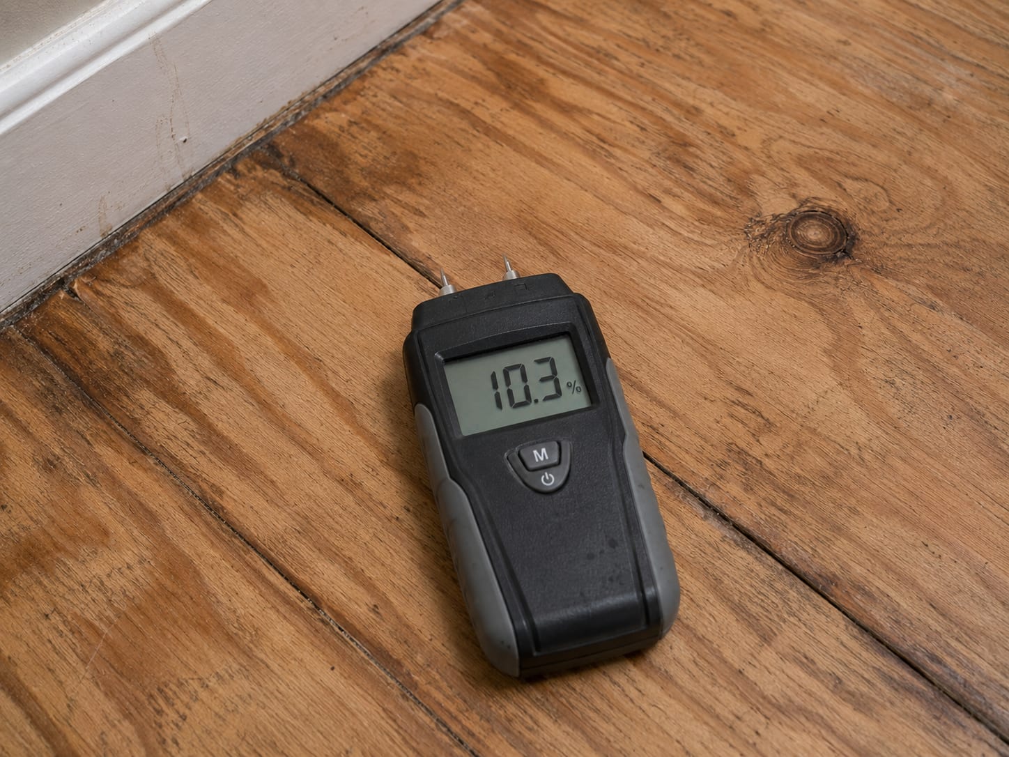 Digital pin-type moisture meter resting on a dried wood subfloor panel showing a low moisture reading after burst pipe restoration