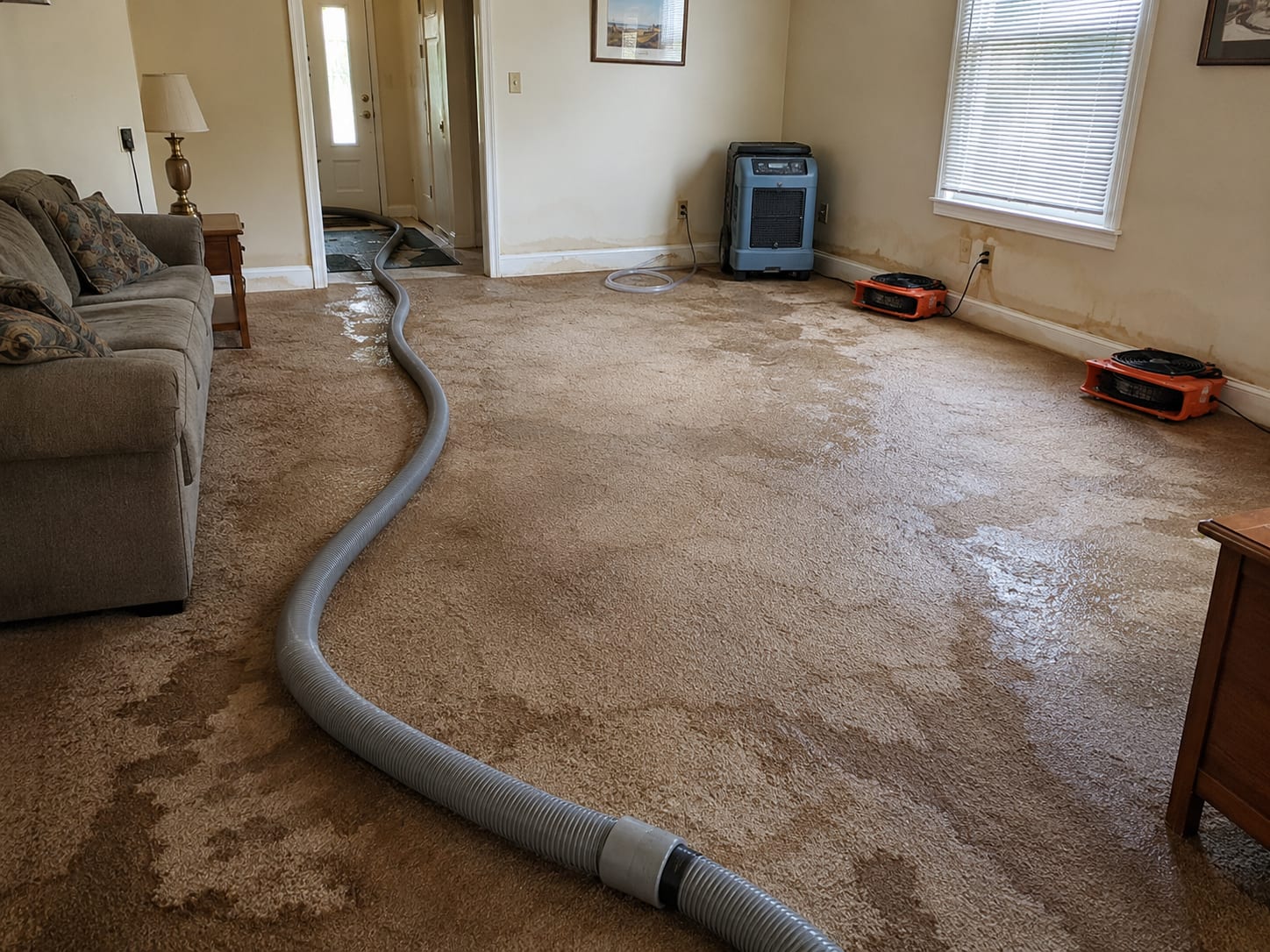 Wide view of a water-damaged residential living room with large extraction equipment on soaked carpet and air movers positioned along baseboards