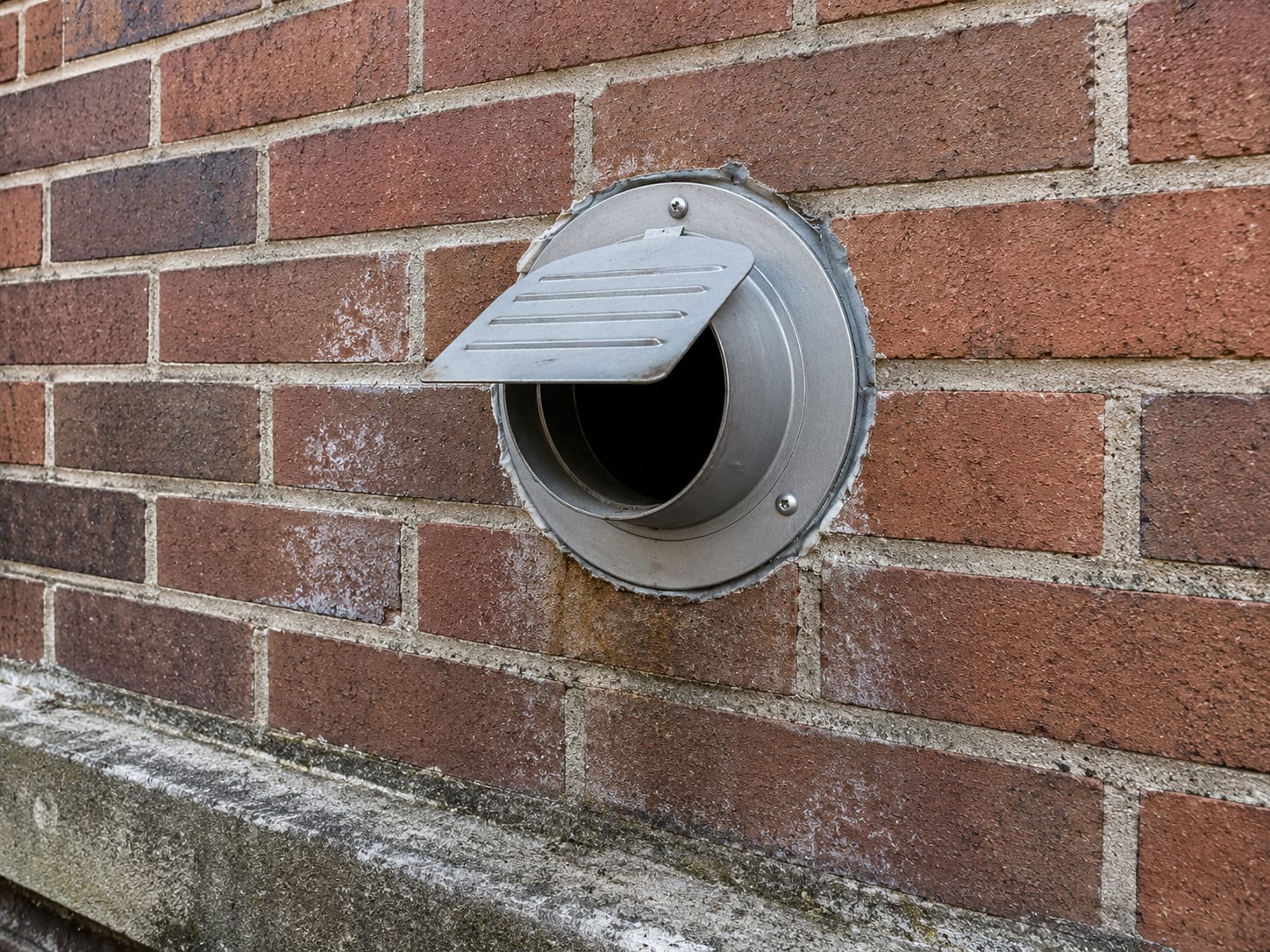 Exterior brick wall of a New Jersey commercial building showing a cleaned wall-mounted dryer vent cap with the louvered flap open and no lint obstruction, surrounded by weathered mortar joints