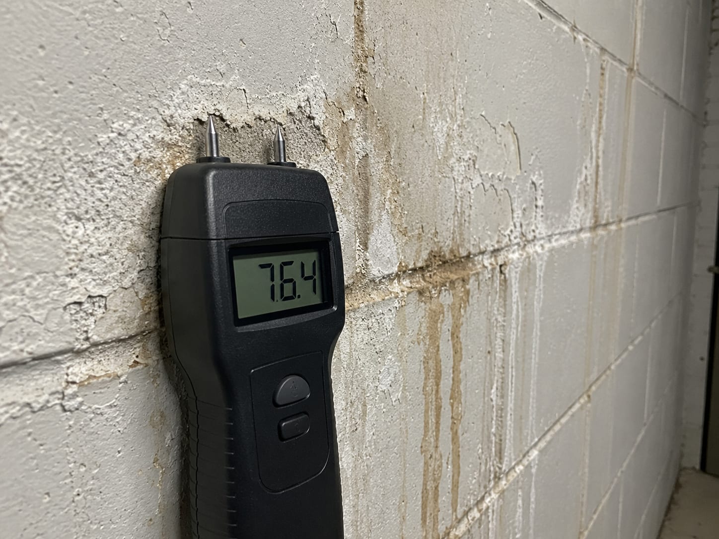 Digital moisture meter probe pressed against a painted concrete block wall in a commercial basement showing elevated moisture reading on the display, surrounded by visible efflorescence and water staining
