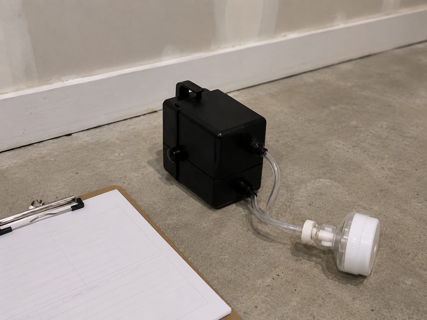 Post-remediation air quality sampling pump with a clear cassette attachment resting on a clean concrete basement floor beside a clipboard with paper forms