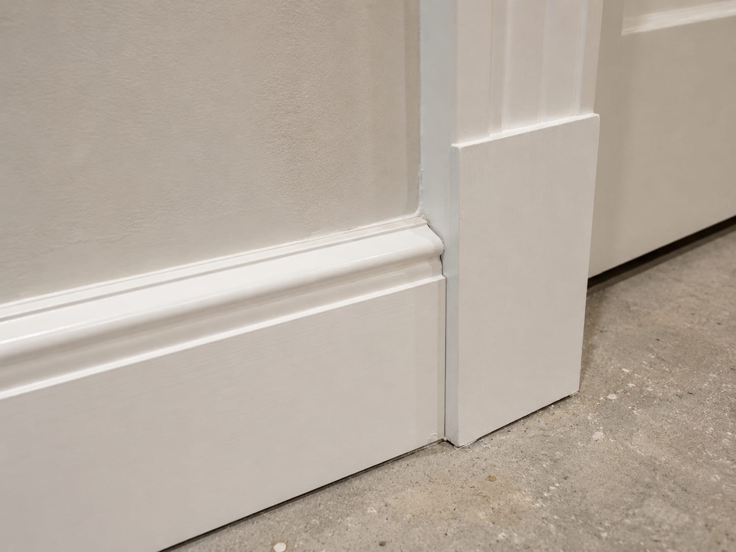 Close-up of a precisely mitered inside corner where new painted baseboard trim meets painted door casing on a freshly drywalled basement wall
