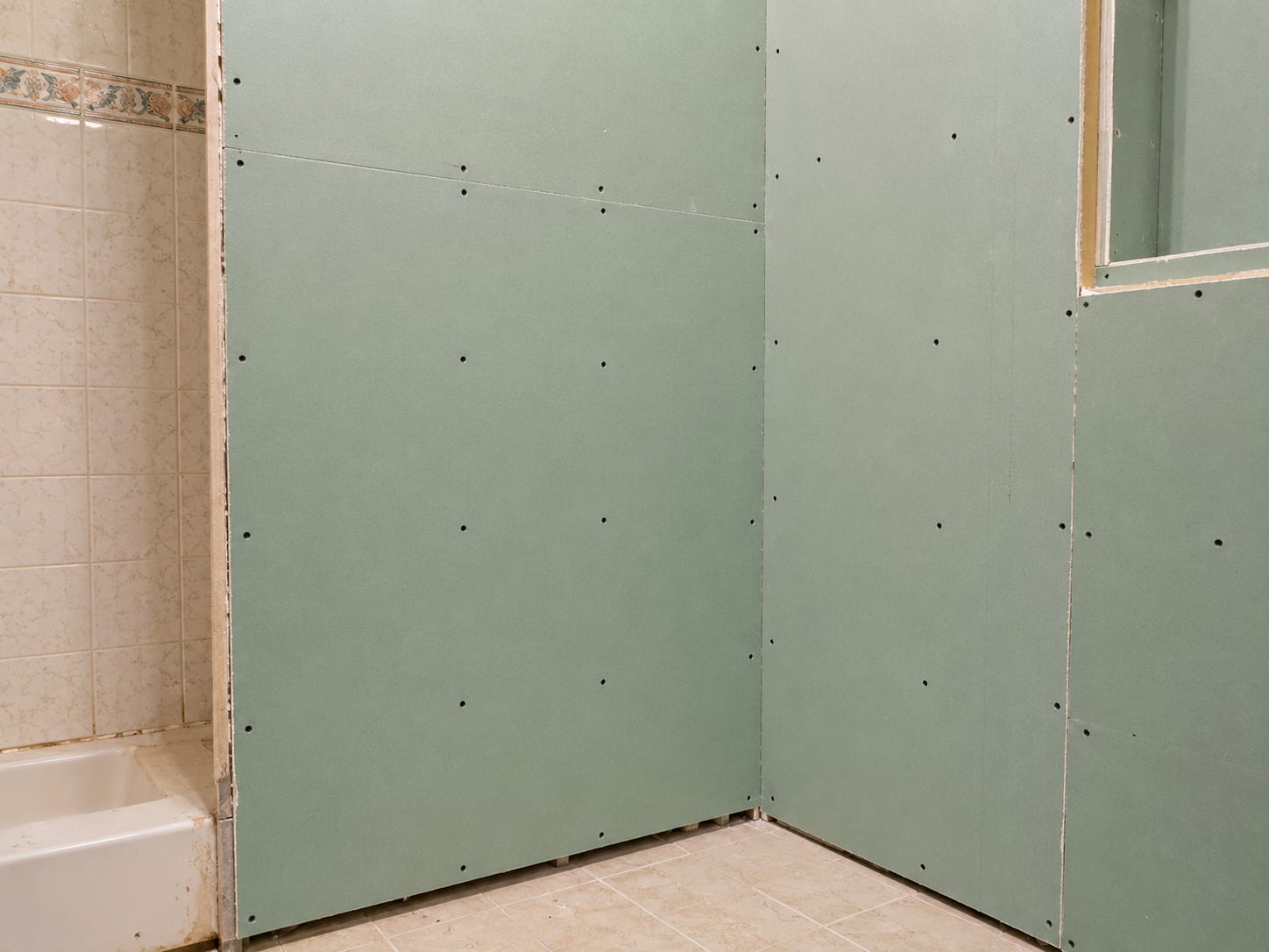New drywall panels screwed into wall studs in a small bathroom of a New Jersey home, moisture resistant green board visible, screw dimples unfilled, raw edges near floor