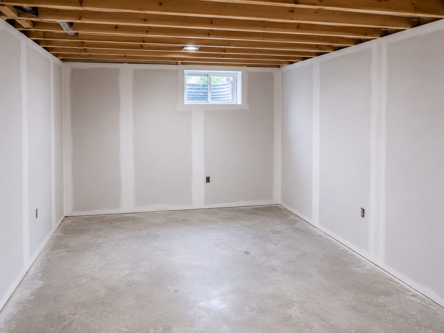 Freshly installed and mudded drywall in a finished basement room of a New Jersey colonial home, clean white walls with smooth joint compound seams visible, natural light entering from a small egress window