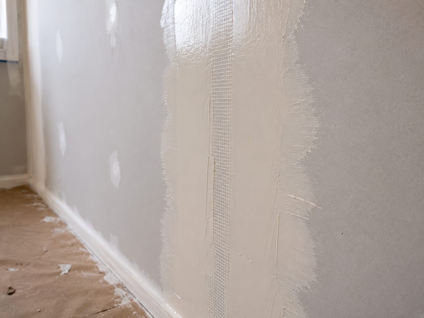 Wide drywall seam with fresh joint compound applied in a smooth feathered coat along a wall in a New Jersey home interior, butt joint edges and mesh tape visible beneath wet compound