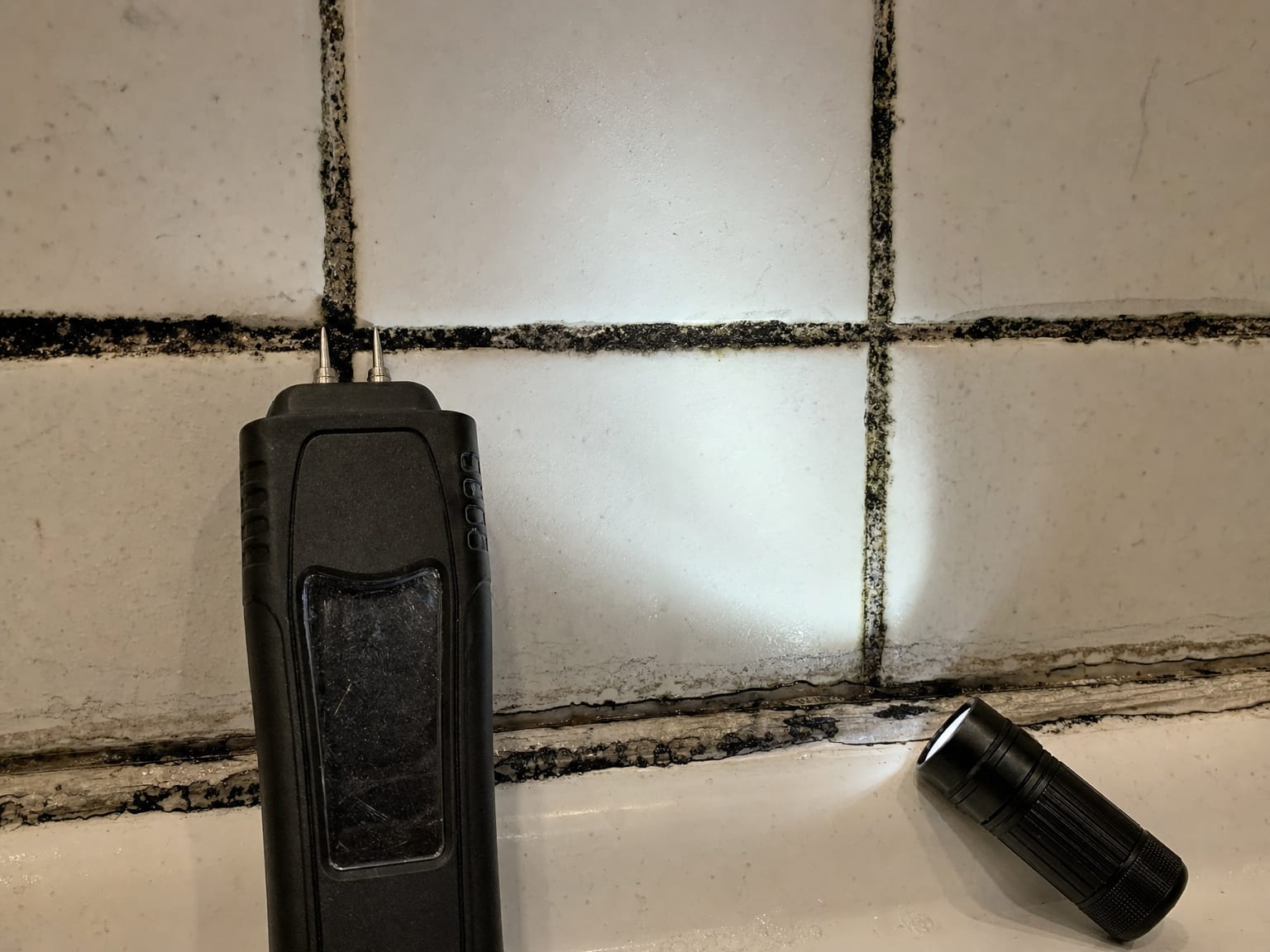 Close-up of bathroom tile grout lines with black mold growth and a moisture meter probe resting against the tile near a caulk seam