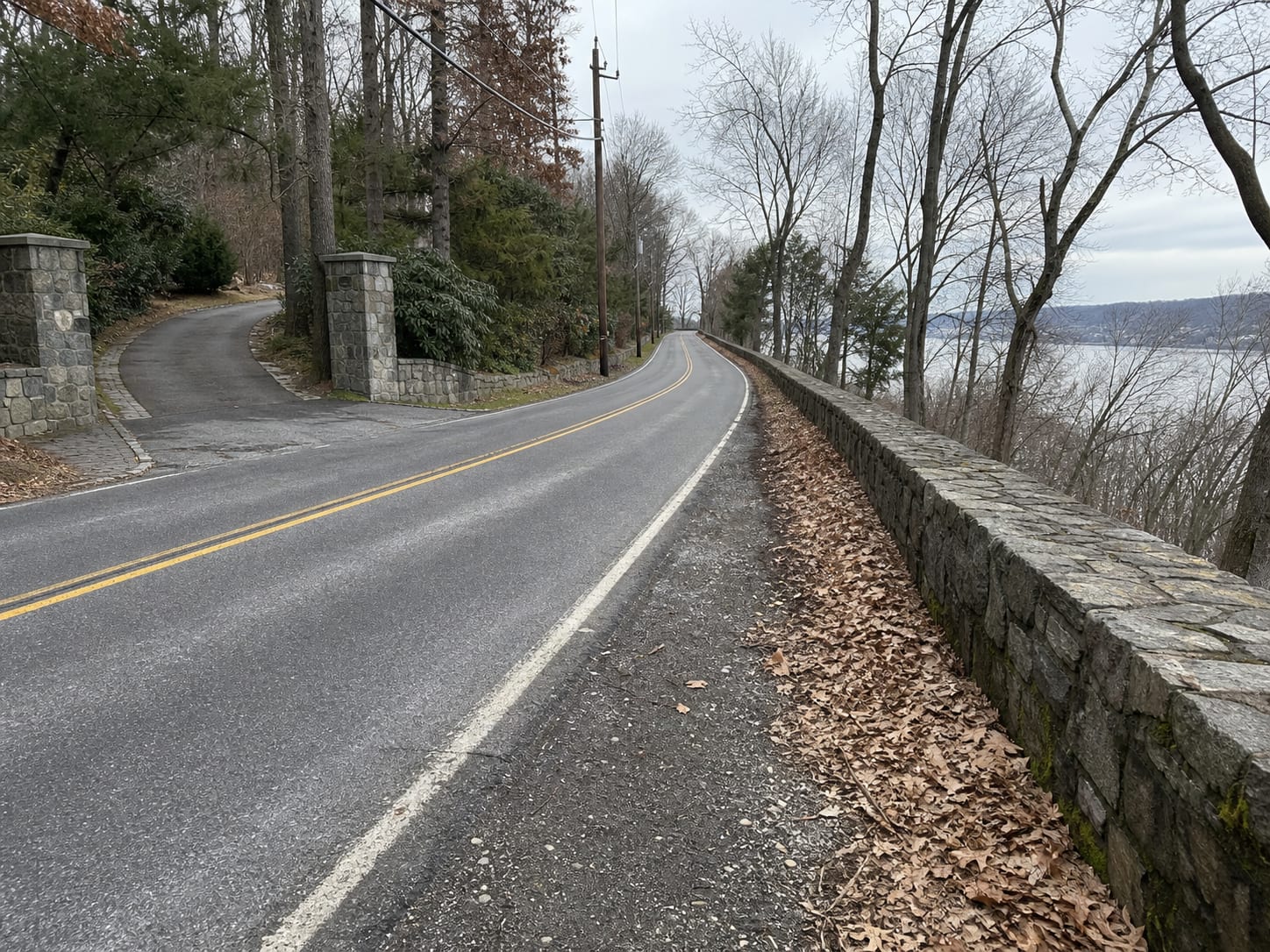 A two lane road along the Palisades ridge in northeastern Bergen County, bordered by dense hardwood woodland, a low stone parapet wall and mortared gate piers at a private drive