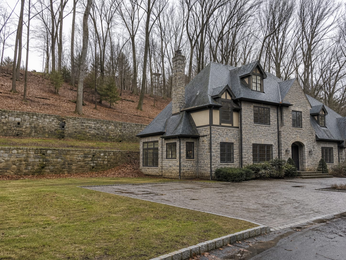 A large detached stone and stucco house set back on a wooded lot in northeastern Bergen County, with ground rising behind it toward a mortared stone retaining wall