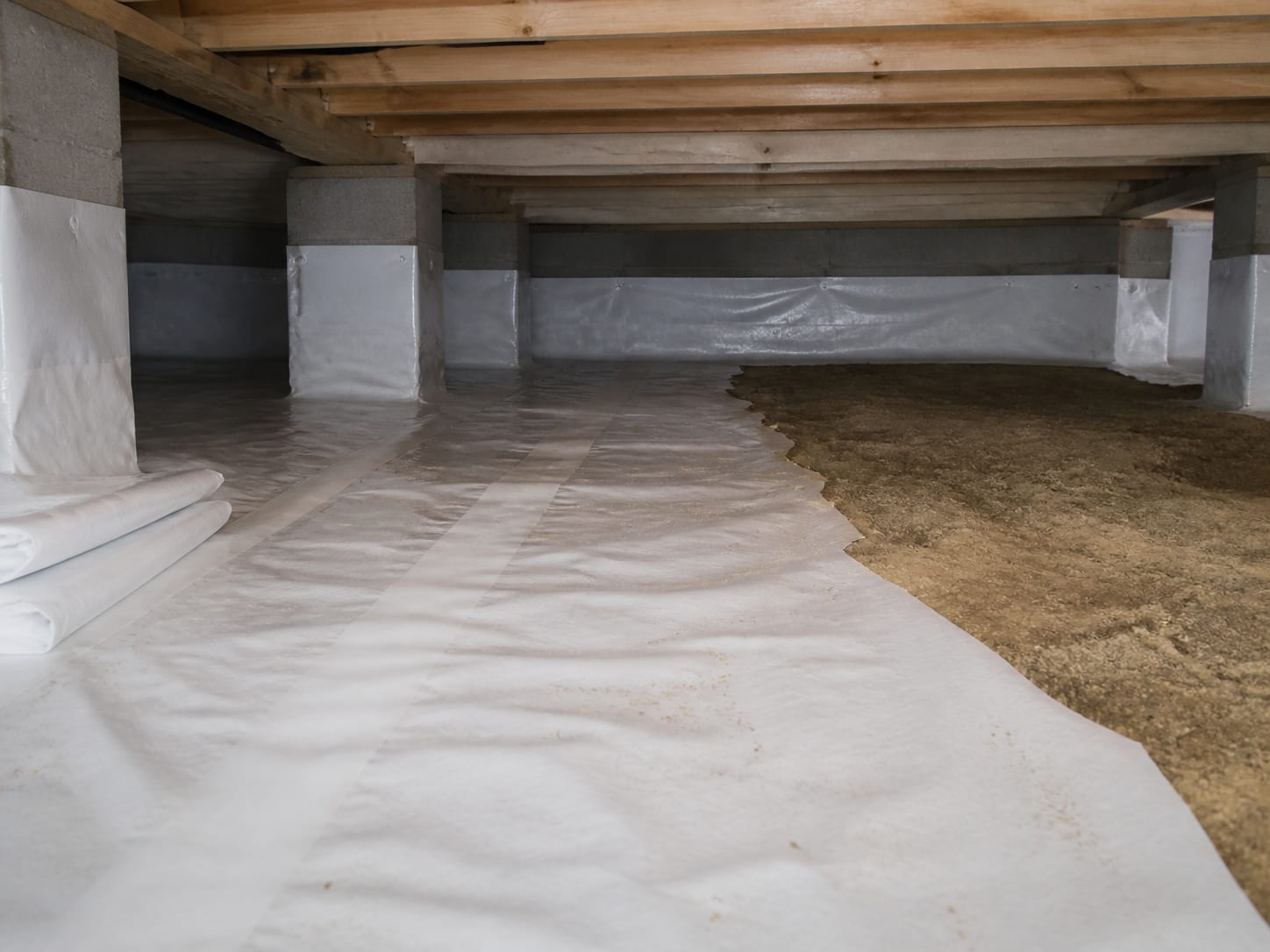 New white vapour barrier being laid across the sandy soil floor of a crawl space in Barnegat, New Jersey, with seams taped and the sheeting run up the block piers