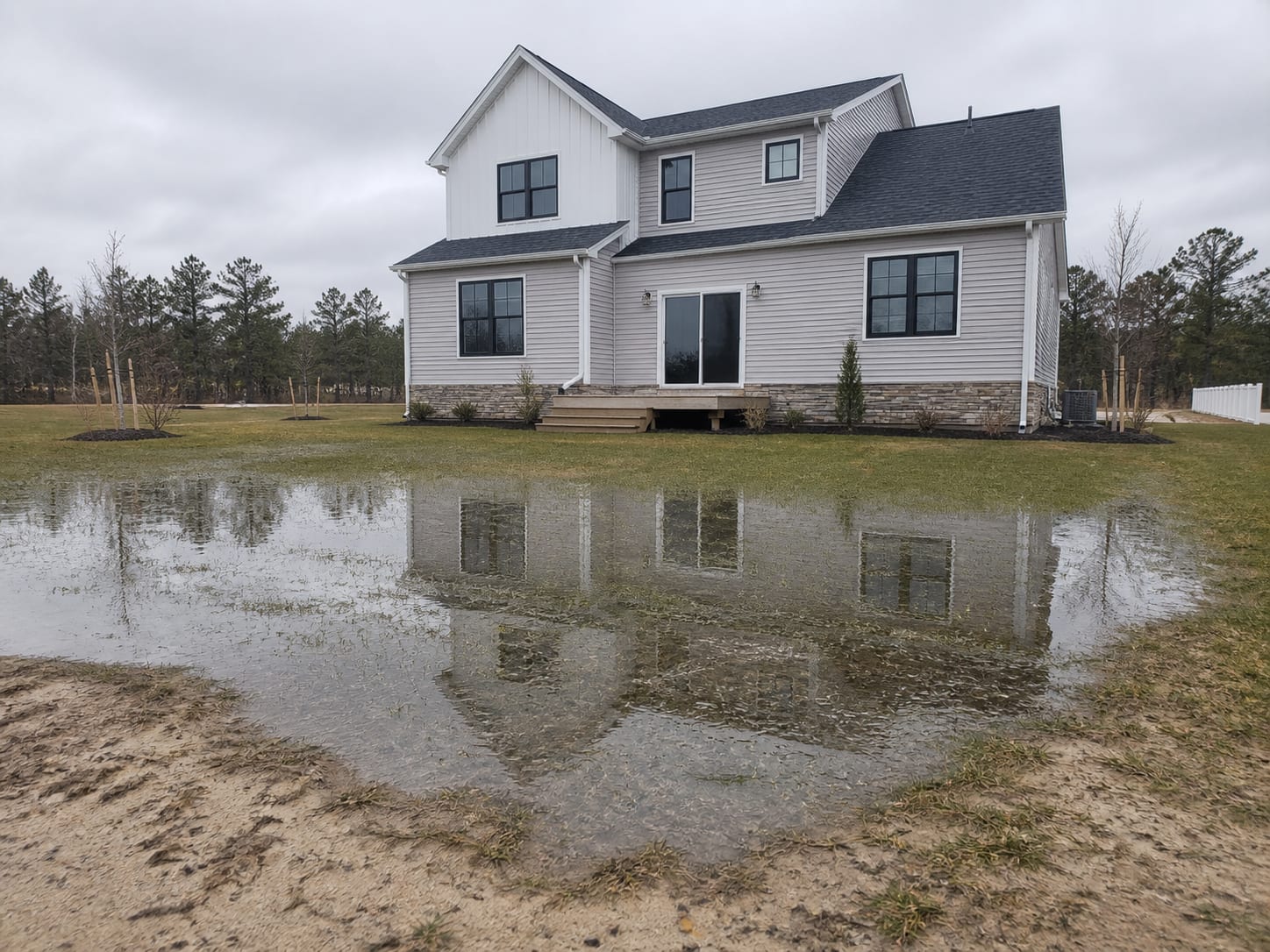 A shallow sheet of standing water lying across the flat young rear lawn of a recently built detached house in Barnegat, New Jersey, with thin new landscaping and open sky behind