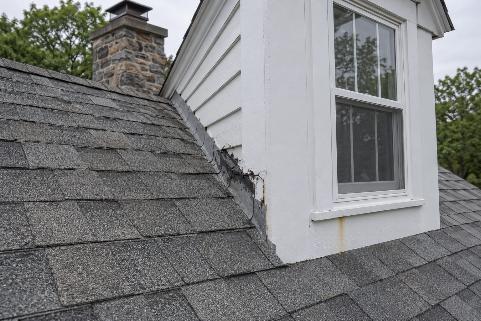 The corner of a dormer window on a roof with an old tar patch over a flashing seam and a faint streak of staining on the siding below, on a house in Belle Mead, New Jersey