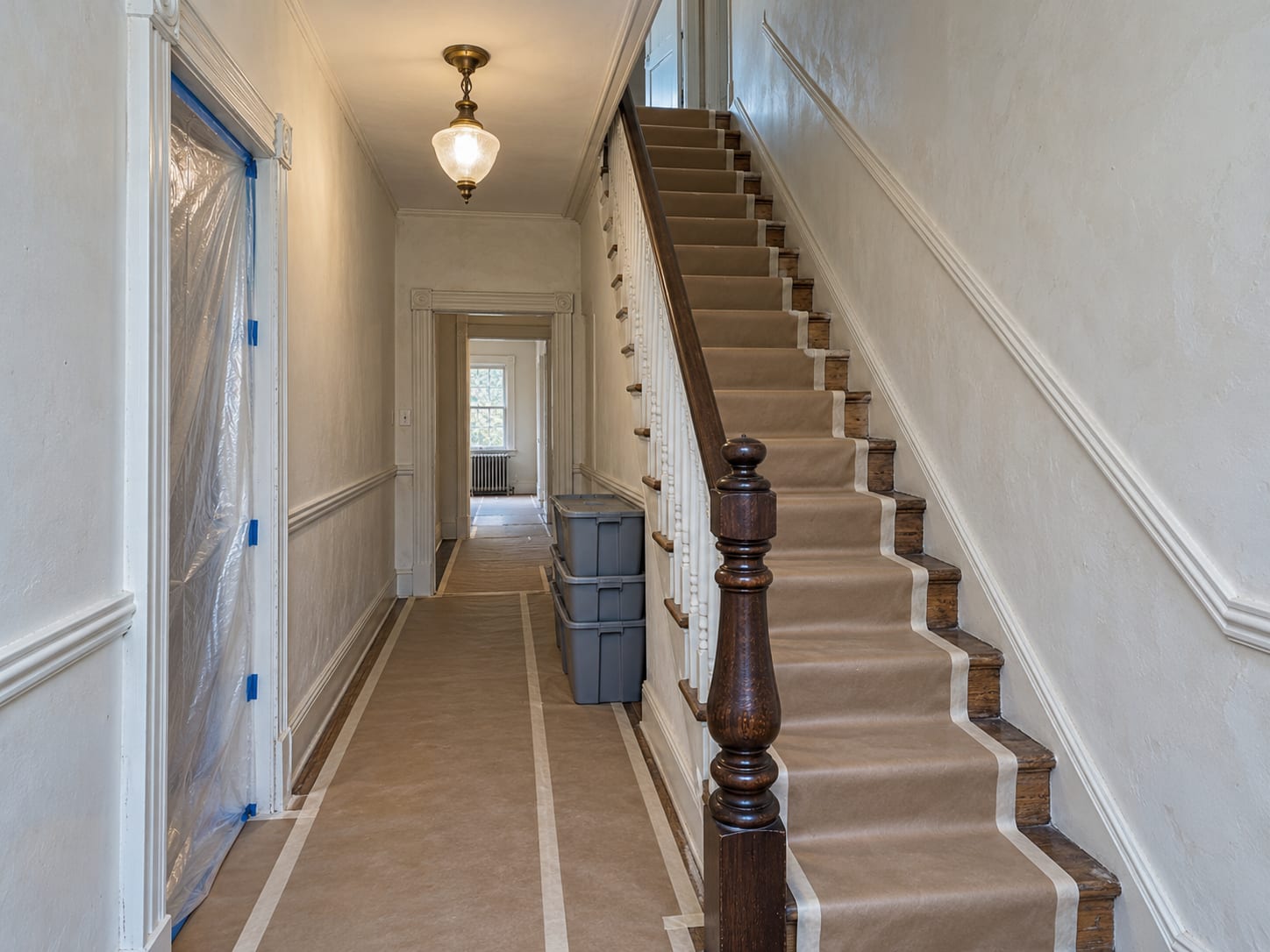 A steep narrow stair hall in an old row house with brown paper floor protection laid up the steps to a doorway covered with taped plastic sheeting, in Bordentown, New Jersey