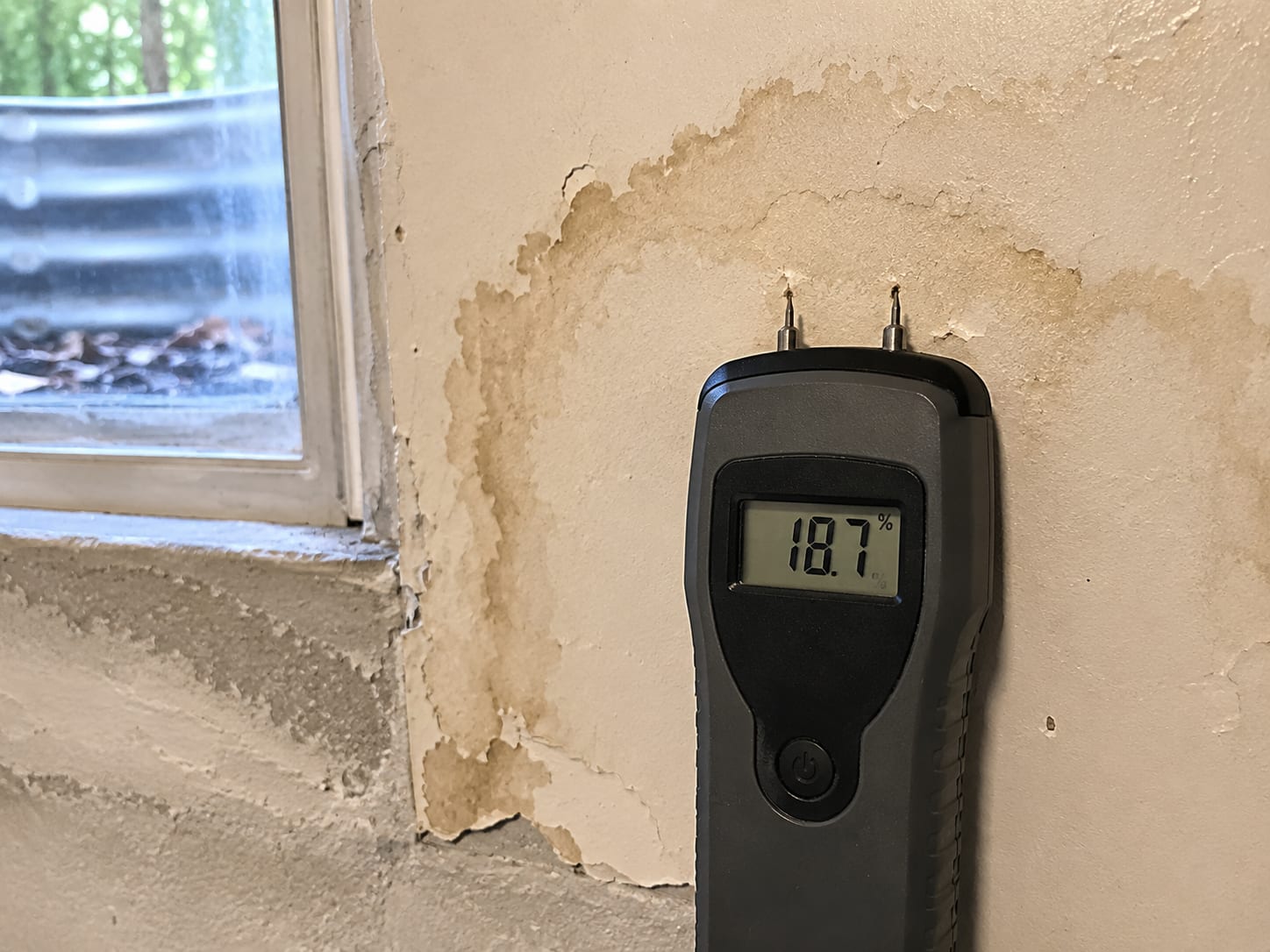 Digital moisture meter with pin probes pressed into a water-stained drywall surface near a basement window well, moisture reading visible on the small display