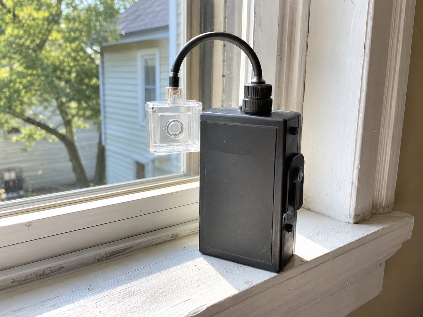 Air sampling pump with a cassette filter attached resting on a windowsill inside a residential room with natural daylight coming through the window