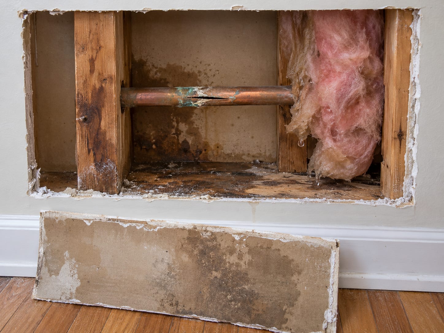 A wall cavity opened at the bottom, showing a split copper pipe with green corrosion, a water stained stud and a cut piece of wet drywall, in a Franklin Township, New Jersey house