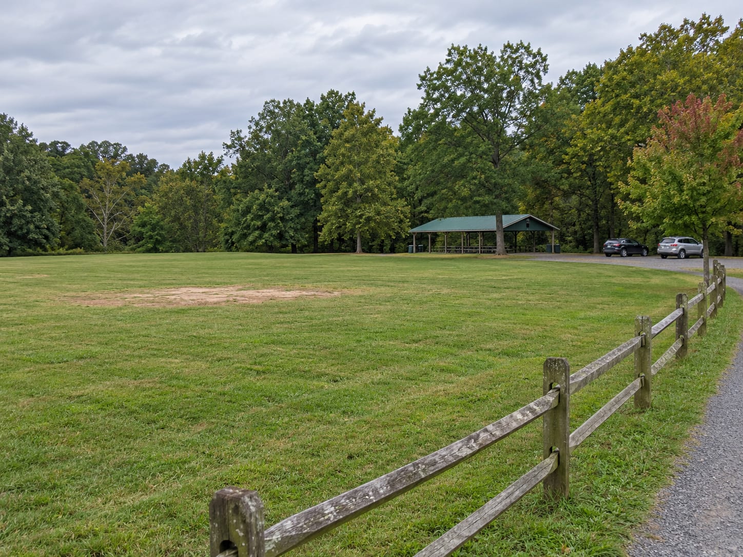 Open mown playing fields beside a gravel path and a wooded edge, with a split rail fence and a small parking area, in a park in Franklin Township, New Jersey