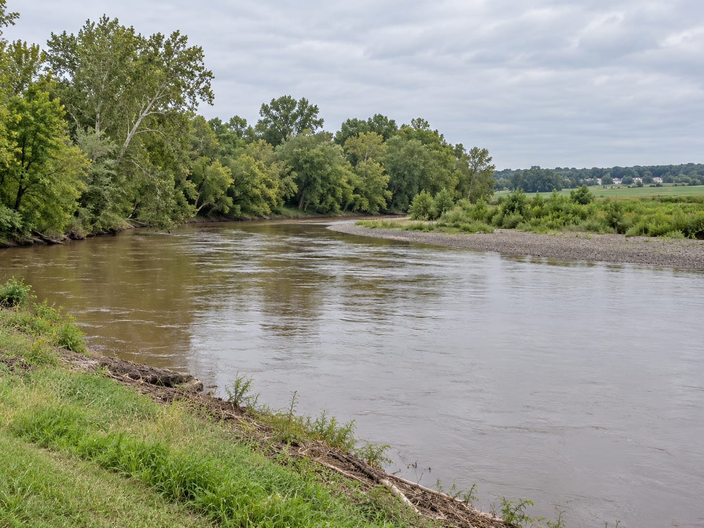 A broad slow river flowing past a gravel bar and a wide wooded floodplain with farm fields beyond, in Franklin Township, New Jersey
