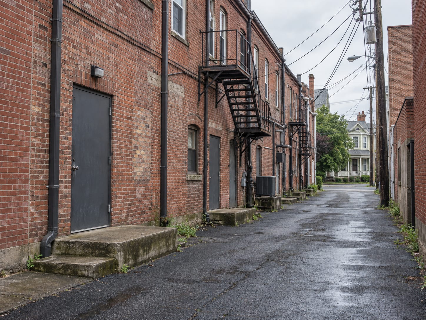 A narrow rear service lane runs behind a brick commercial row in downtown Hightstown, with steel back doors and apartment windows above.