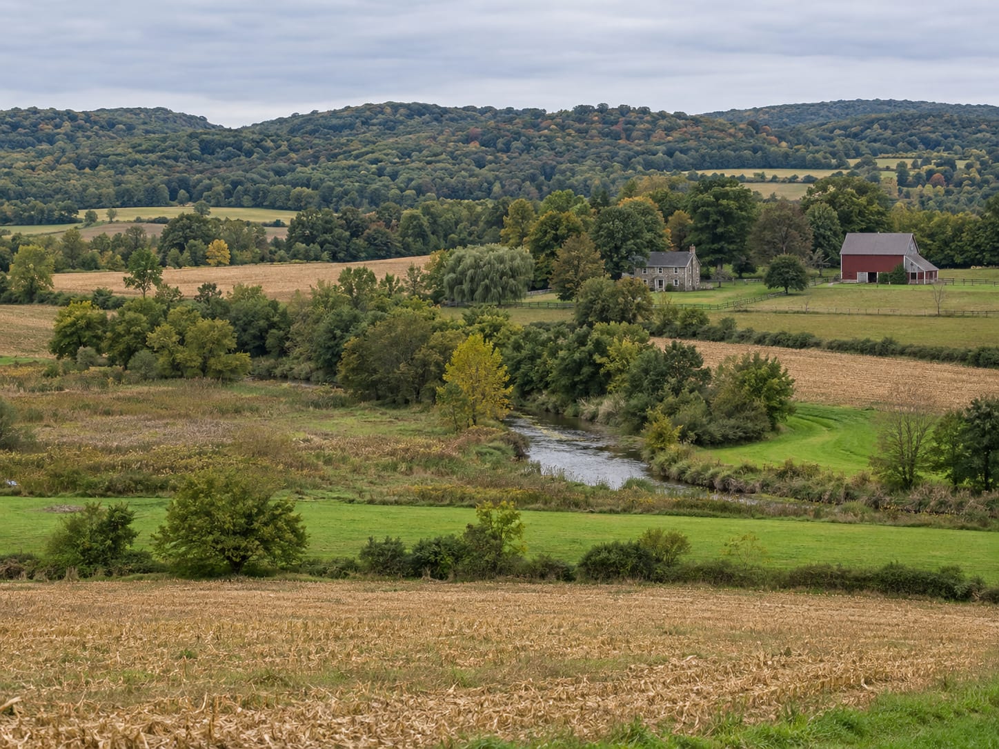A broad valley of farm fields and hedgerows with a creek winding through a wooded strip toward low hills, in Hopewell, New Jersey