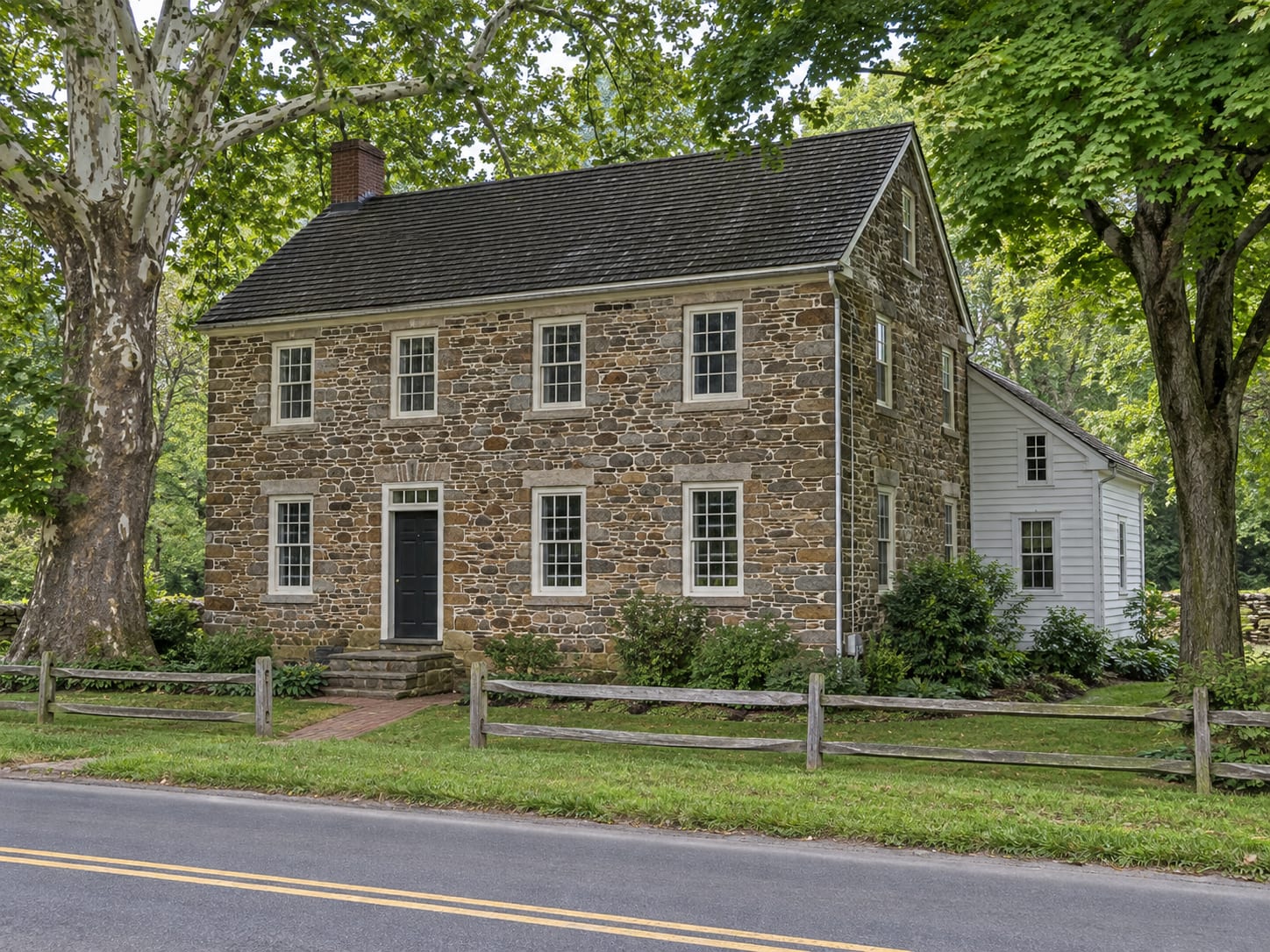 An eighteenth or early nineteenth century two storey house of coursed fieldstone with a later white clapboard wing, standing close to a two lane road under mature trees, in Kingston, New Jersey
