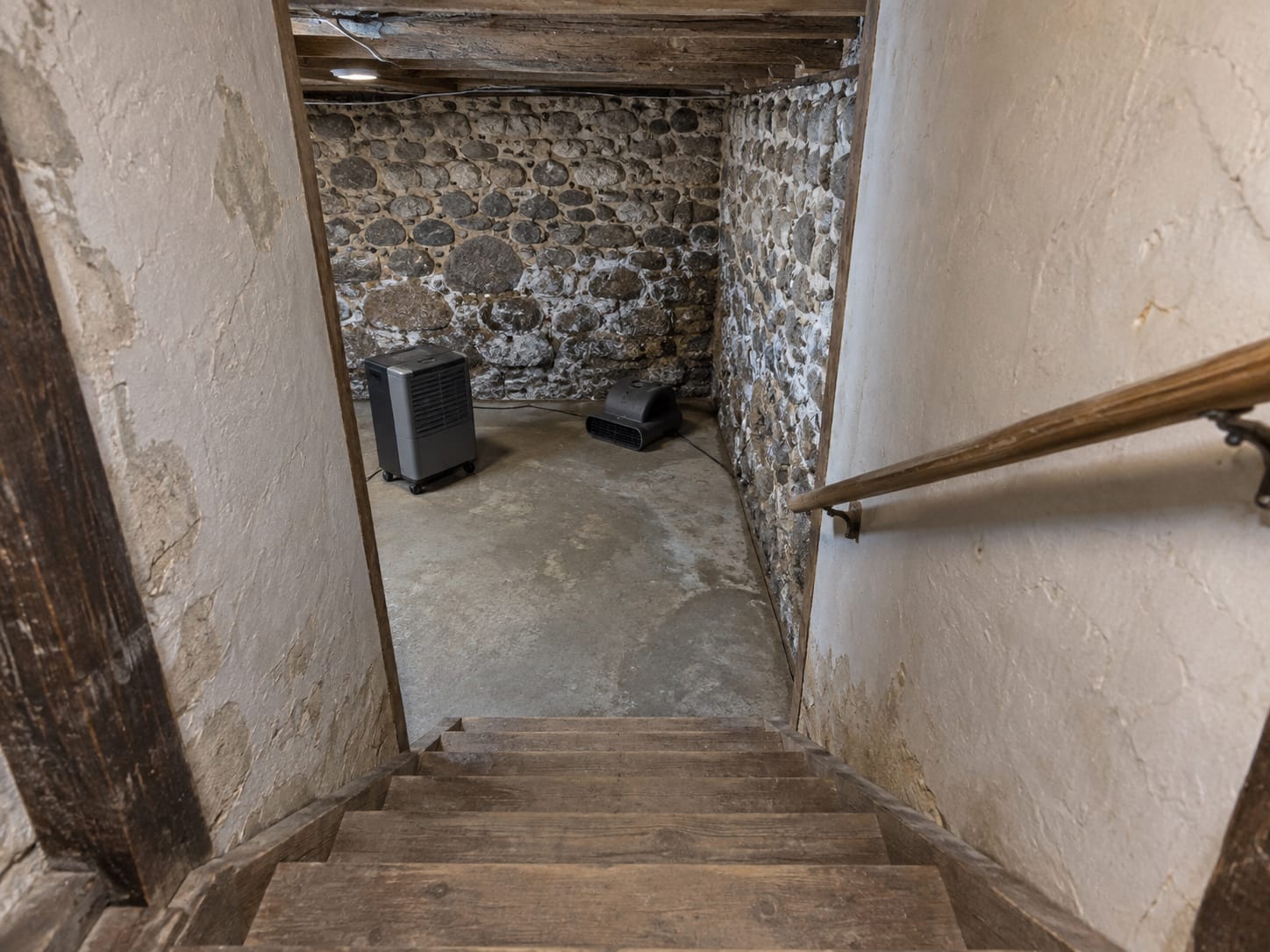 A steep narrow wooden stair leading down into a fieldstone walled cellar in an older Kingston, New Jersey house, with a dehumidifier and air mover drying the floor below