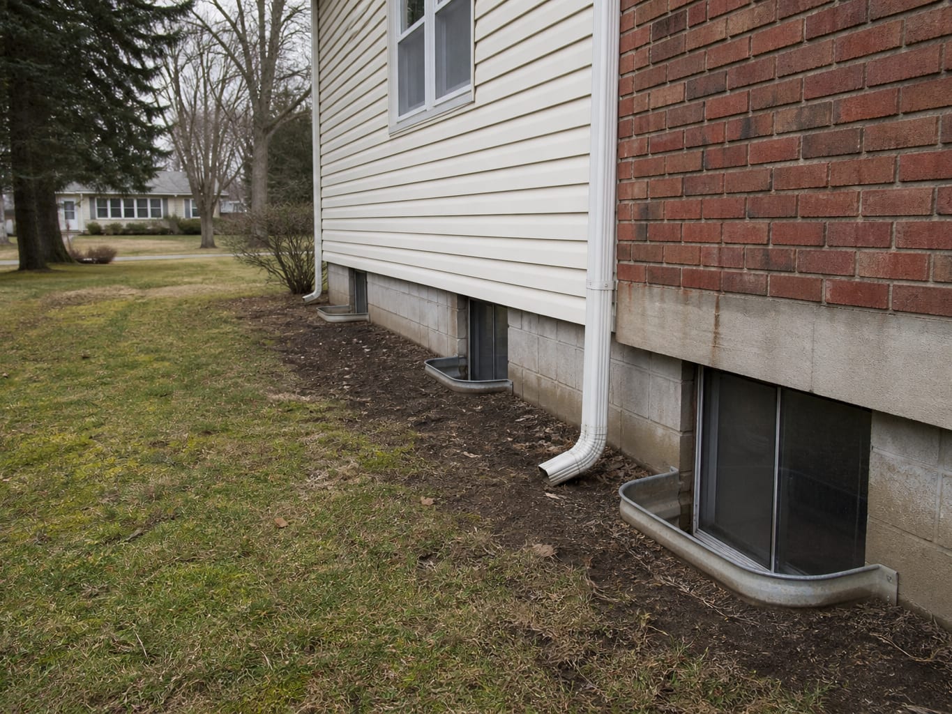 Exterior of a mid-century split-level home in inland New Jersey where a downspout discharges beside a basement window well at the foundation