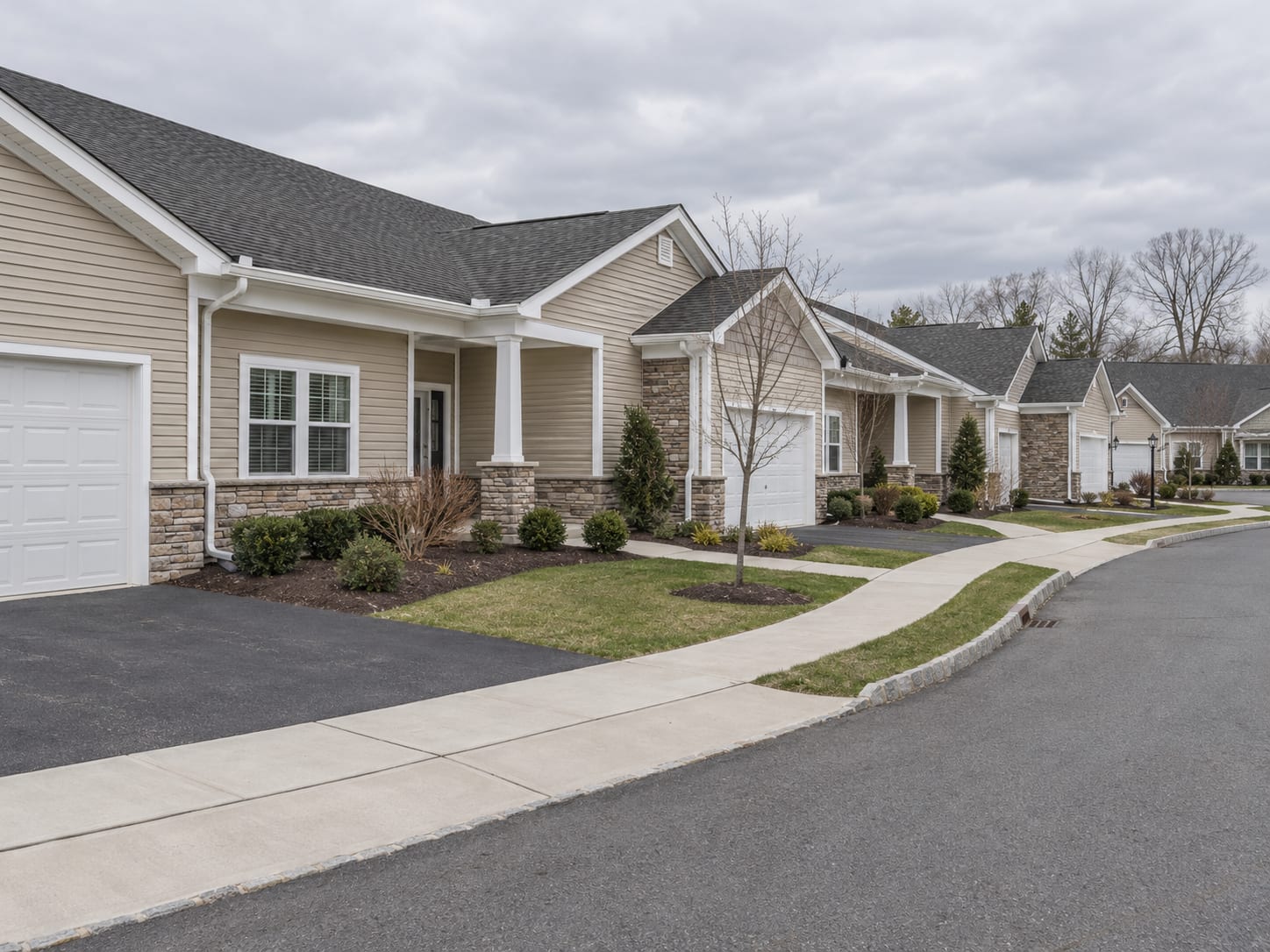 A cluster of single storey attached patio homes in beige vinyl with stone veneer, shared rooflines, single garages and tidy mulch beds, in Monroe Township, New Jersey