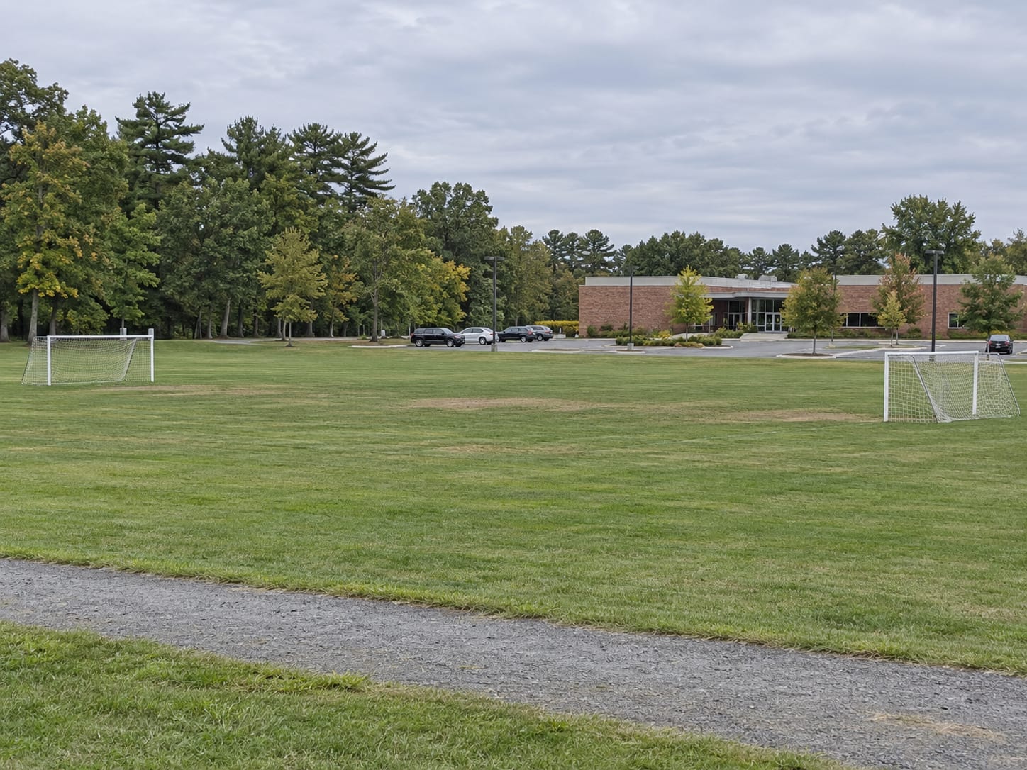 Open mown athletic fields with goalposts beside a gravel path and a treeline, and a low brick library building beyond a parking lot, in Monroe Township, New Jersey