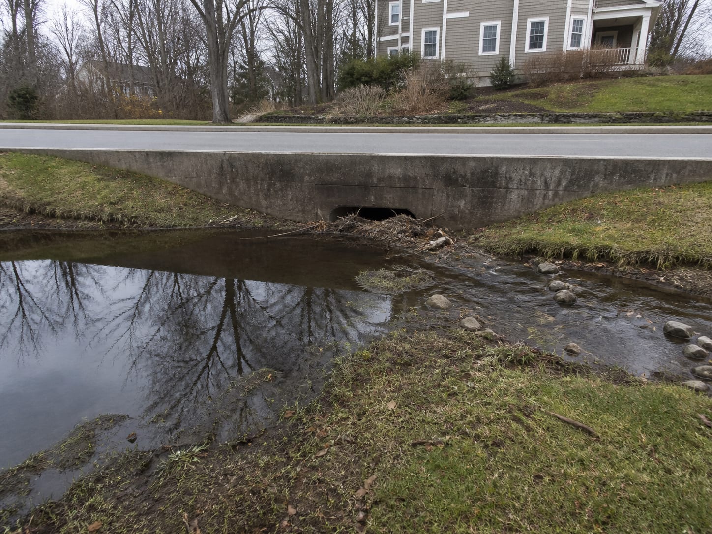 A brook backing up at an undersized culvert headwall beneath a residential street in Montclair, New Jersey, with debris racked against a pipe much narrower than the channel feeding it