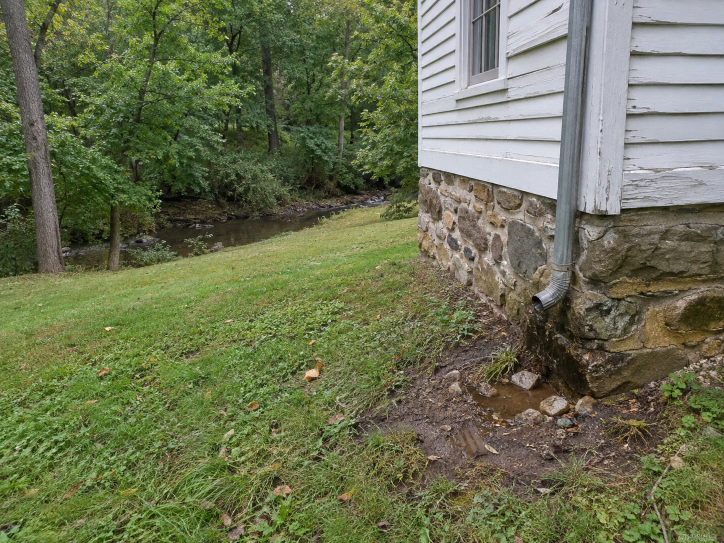 A downspout ending at the base of a fieldstone foundation on bare wet soil, with a small stream and wooded bank behind an older house, in Montgomery, New Jersey