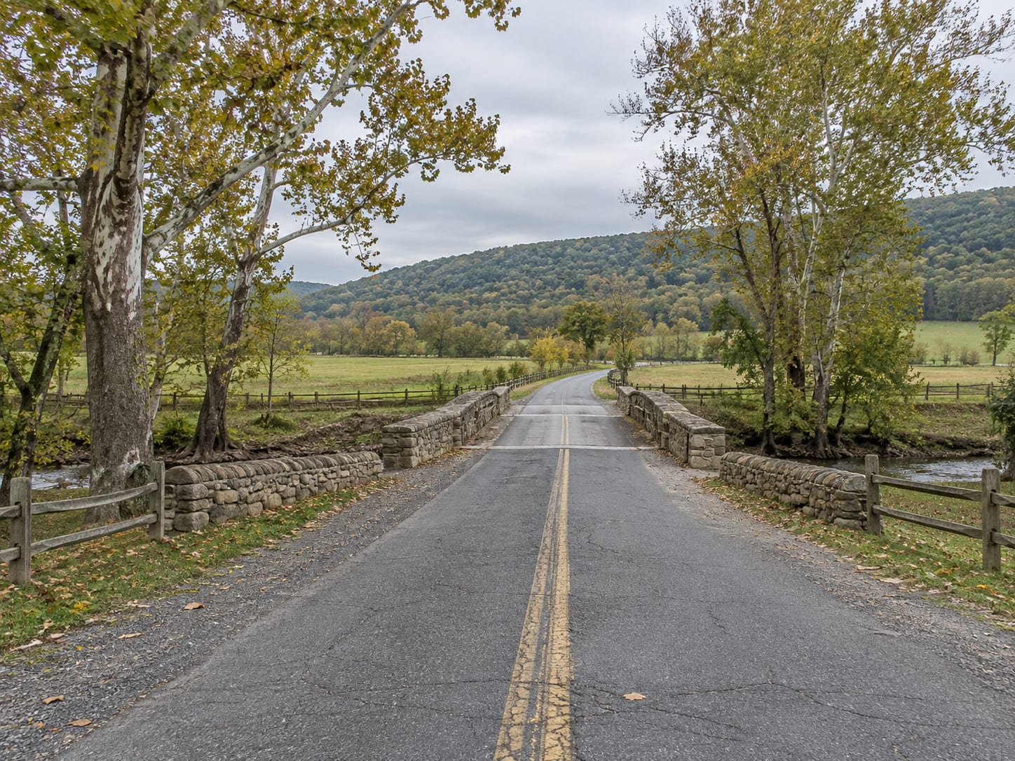 A two lane country road crossing a small stone arched bridge over a stream, with farm fields, a stone wall and a wooded ridge beyond, near Montgomery, New Jersey