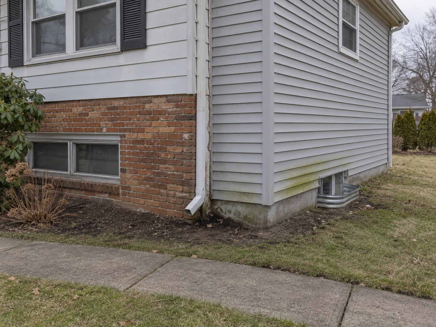 The vertical seam where a vinyl sided addition meets an older brick and siding house, with a downspout emptying at the corner and staining on the base, in North Brunswick, New Jersey