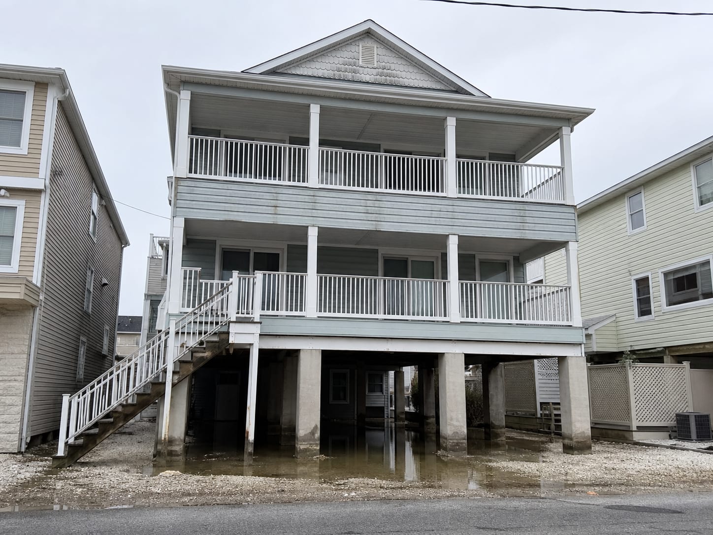 A shore duplex raised on piers above an open parking undercroft in Ocean City, New Jersey, with shallow water standing on the sand beneath it and a tide line marking the piers