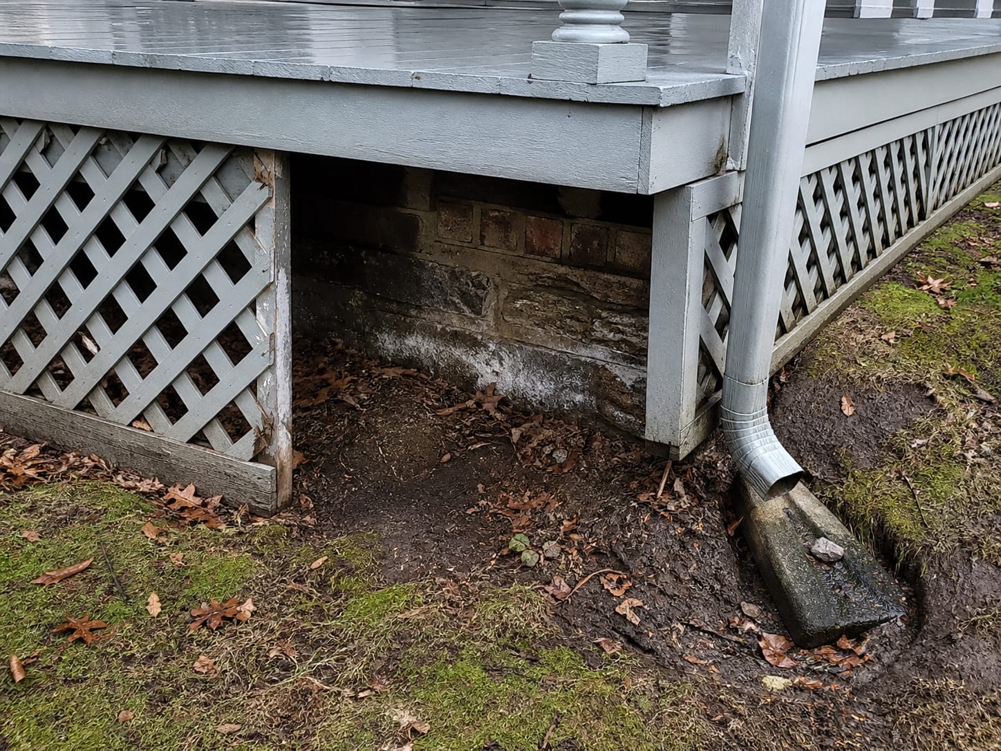 The lattice skirt and stone foundation beneath the wraparound porch of an older frame house in Ridgewood, New Jersey, with efflorescence on the masonry and a downspout discharging alongside